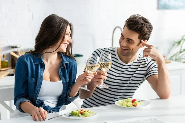 Alegre pareja tintineo vasos de vino cerca de platos con ensalada en la cocina - foto de stock