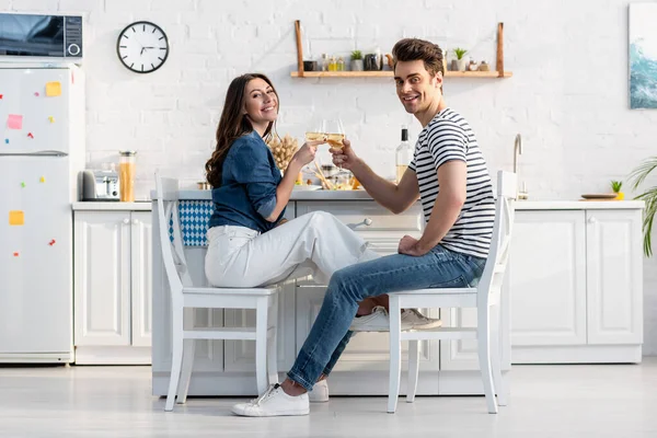 Happy couple holding glasses with wine and clinking while sitting in kitchen — Stock Photo