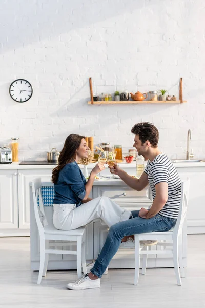 Vista lateral do casal feliz copos clinking com vinho na cozinha — Fotografia de Stock