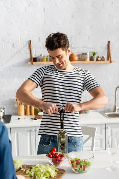 Homem feliz sorrindo ao abrir garrafa com vinho na cozinha — Fotografia de Stock