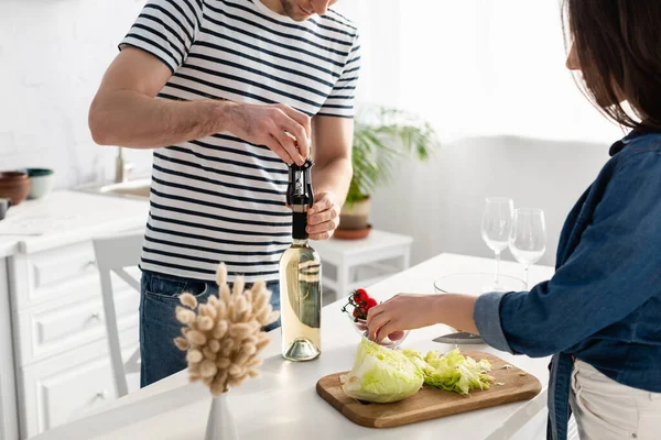 Donna taglio lattuga vicino fidanzato aprendo bottiglia di vino — Foto stock