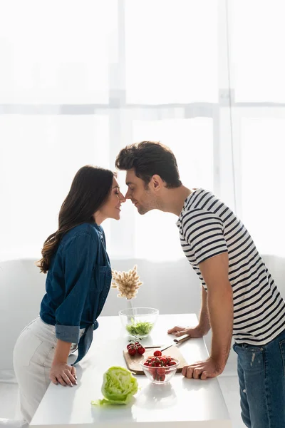 Vista lateral de feliz pareja con los ojos cerrados tocando las narices cerca de los ingredientes en la mesa - foto de stock