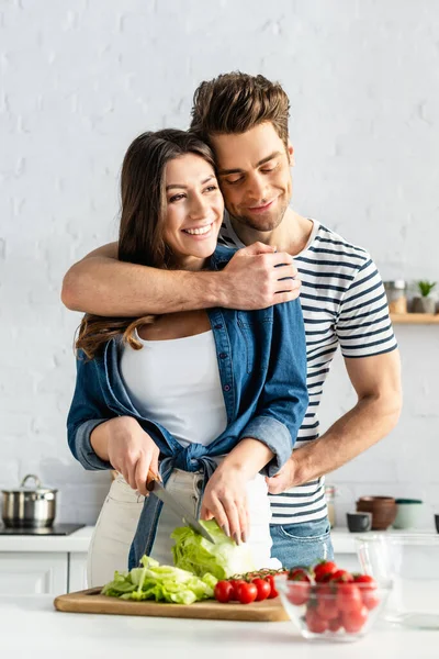Happy man hugging woman preparing salad in kitchen — Stock Photo