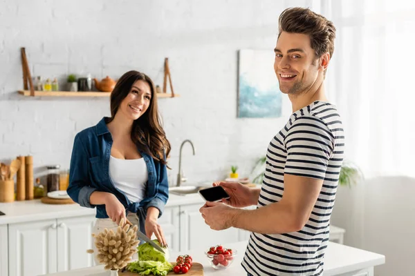 Щасливий чоловік тримає смартфон з порожнім екраном, коли жінка готує на кухні — стокове фото