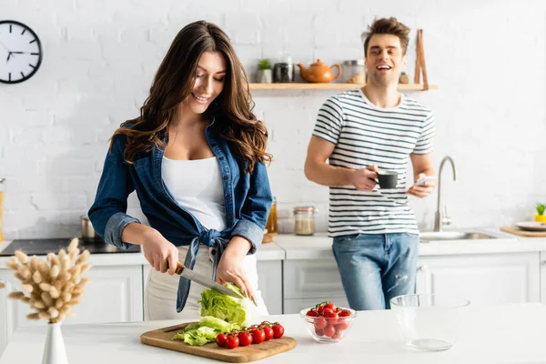 Fröhliche Frau kocht, während Mann Tasse und Smartphone auf verschwommenem Hintergrund hält — Stockfoto