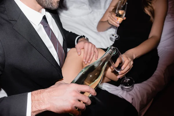 Vista ritagliata dell'uomo in giacca e cravatta versare champagne in vetro vicino fidanzata sul letto su sfondo sfocato in hotel — Foto stock