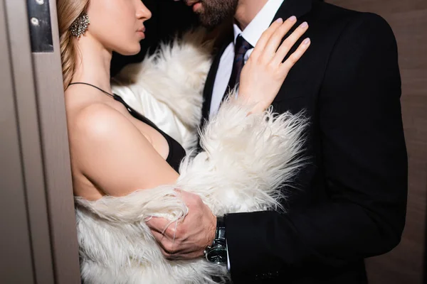 Cropped view of woman in fluffy jacket hugging boyfriend in suit in hotel during night — Stock Photo