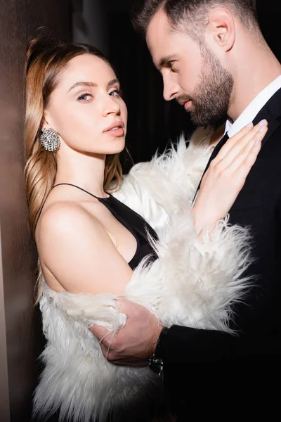 Woman in fluffy jacket looking at camera while embracing boyfriend in suit in hotel — Stock Photo