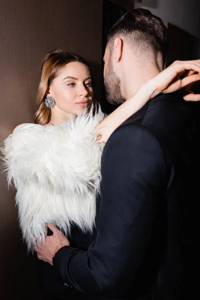 Mulher elegante em casaco fofo abraçando namorado em terno no hotel durante a noite — Fotografia de Stock