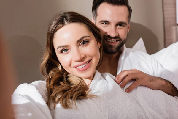 Sorrindo mulher olhando para a câmera perto namorado em roupão de banho no fundo embaçado no hotel — Fotografia de Stock