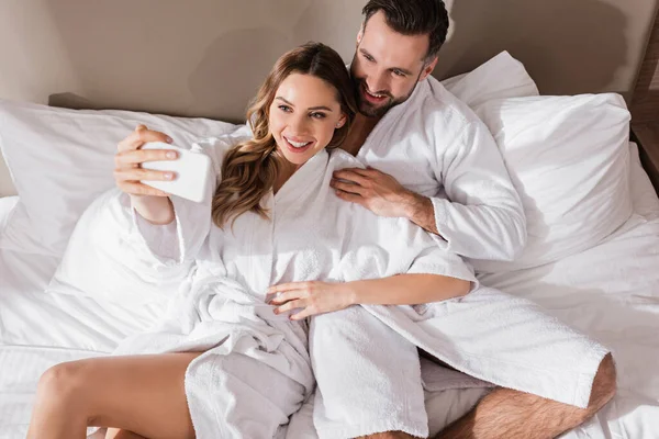 Вид на веселую пару в халатах, делающую селфи со смартфоном на кровати в отеле — стоковое фото