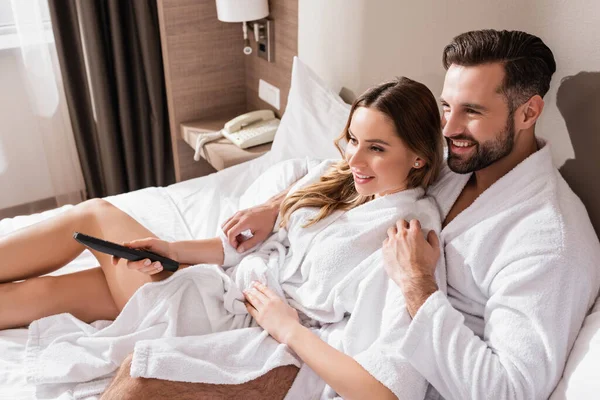Couple in bathrobes hugging while watching tv on bed in hotel — Stock Photo
