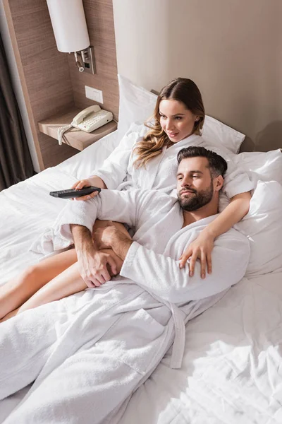 Woman embracing boyfriend in bathrobe and holding remote controller on bed in hotel — Stock Photo