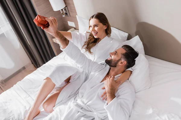 Homem alegre em roupão de banho segurando presente perto da namorada na cama do hotel — Fotografia de Stock