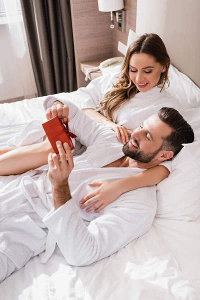 Mann im Bademantel hält Geschenk neben lächelnder Freundin auf Hotelbett — Stockfoto