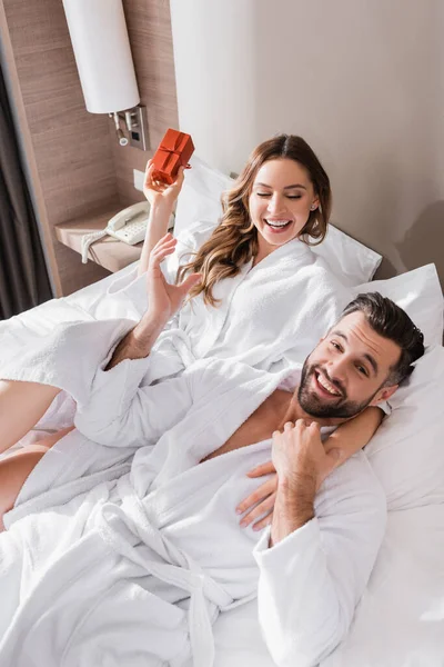 Mujer alegre en albornoz sosteniendo caja de regalo y abrazando novio en la cama del hotel - foto de stock