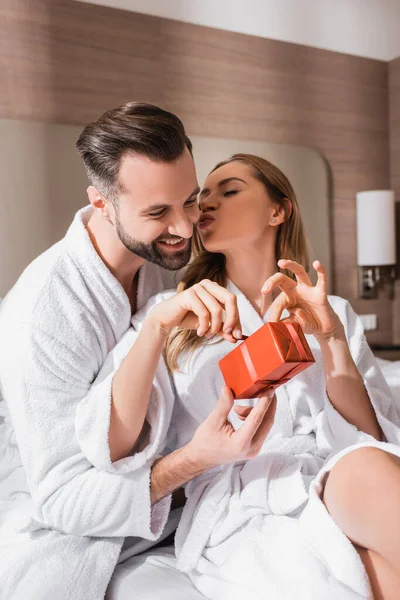 Mulher de roupão beijando namorado enquanto desembalando presente na cama do hotel — Fotografia de Stock