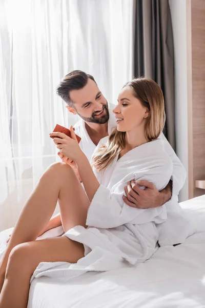 Homme souriant embrassant petite amie avec boîte cadeau en peignoir blanc sur le lit de l'hôtel — Photo de stock