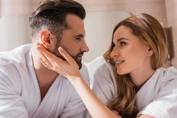 Mujer sonriente en albornoz tocando la cara del novio en la habitación de hotel - foto de stock