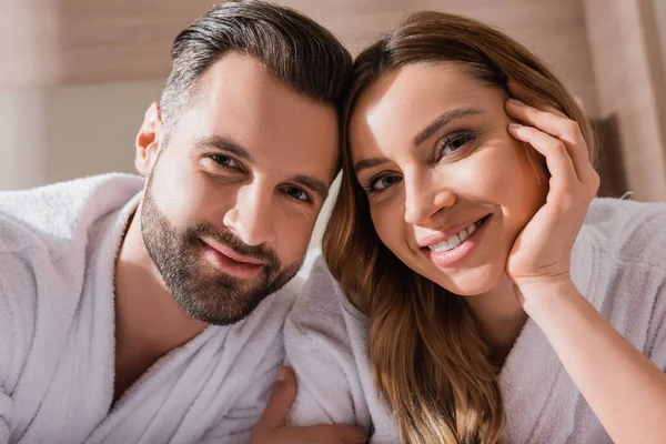 Pareja positiva en albornoces blancos mirando a la cámara en el hotel - foto de stock