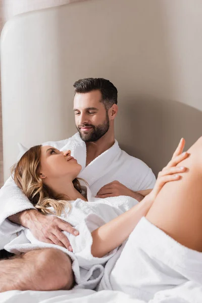 Bearded man embracing woman in bathrobe on white bedding in hotel — Stock Photo