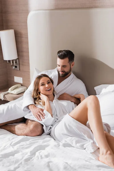 Smiling man hugging girlfriend in bathrobe on bed in hotel room — Stock Photo