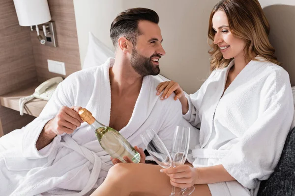 Hombre de albornoz blanco sosteniendo botella de champán cerca de novia sonriente con gafas en la cama del hotel - foto de stock