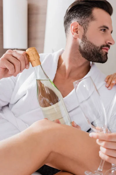 Bottiglia di champagne in mano all'uomo in accappatoio vicino alla fidanzata con bicchieri in primo piano sfocati in hotel — Foto stock