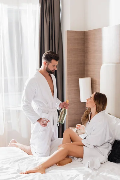 Hombre sosteniendo copas y botella de champán cerca de su novia en albornoz blanco en la cama en el hotel - foto de stock