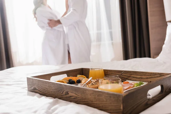 Vista recortada de delicioso desayuno en bandeja en la cama y pareja en albornoces sobre fondo borroso en el hotel - foto de stock