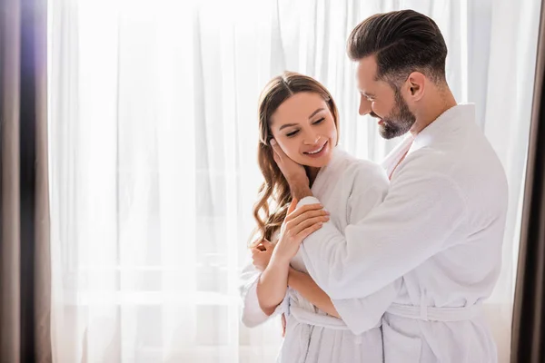 Man in bathrobe touching face of smiling girlfriend near window in hotel room — Stock Photo