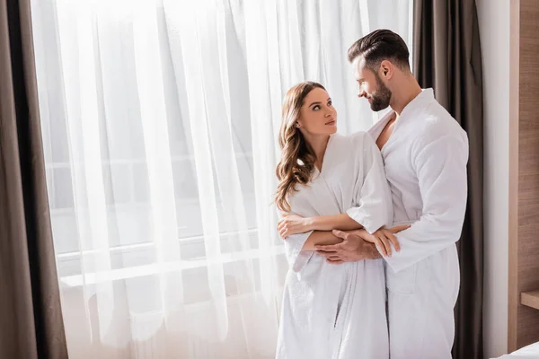 Pareja de albornoces blancos abrazándose cerca de la ventana en la habitación del hotel - foto de stock