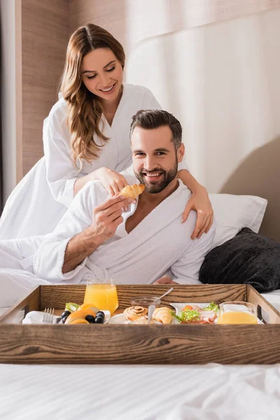 Hombre sonriente sosteniendo croissant cerca de novia en albornoz y desayuno en bandeja en primer plano borroso en el hotel - foto de stock