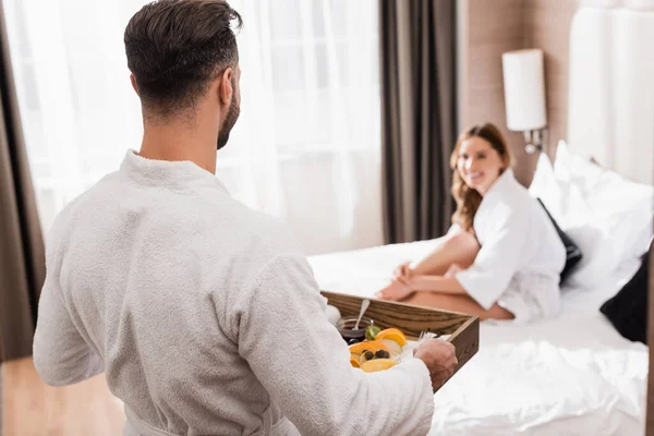 Man in bathrobe holding breakfast tray near girlfriend on hotel bed on blurred background — Stock Photo