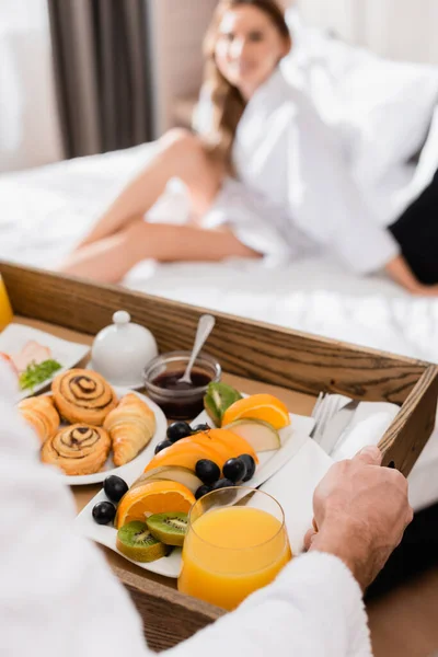 Hombre sosteniendo bandeja de desayuno con zumo de naranja y frutas cerca de la novia en la cama del hotel sobre fondo borroso - foto de stock