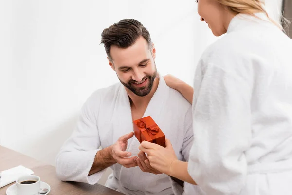 Woman in bathrobe giving present to smiling boyfriend near coffee in hotel room — Stock Photo