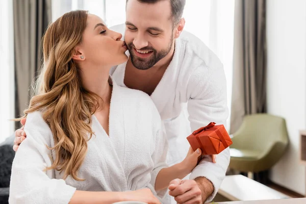 Frau im Bademantel küsst Freund und hält Geschenkbox im Hotelzimmer — Stockfoto