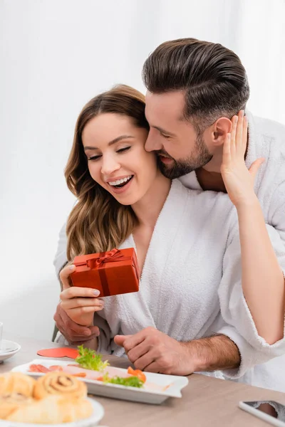 Cheerful woman in bathrobe touching boyfriend while holding gift box near breakfast on blurred foreground in hotel — Stock Photo