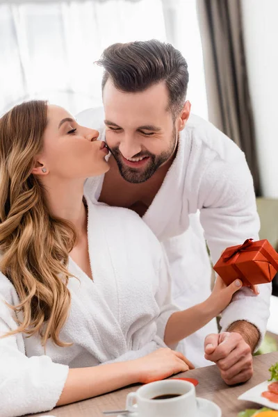 Woman kissing smiling boyfriend and holding present near coffee on blurred foreground in hotel — Stock Photo