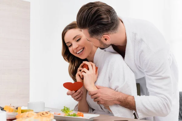 Homme en peignoir embrassant petite amie joyeuse avec cadeau et tenant coeur en papier près du petit déjeuner sur le premier plan flou à l'hôtel — Photo de stock