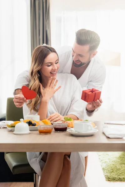 Lächelnder Mann mit Geschenk und Papierherz neben Freundin im Bademantel und Frühstück im verschwommenen Vordergrund im Hotel — Stockfoto