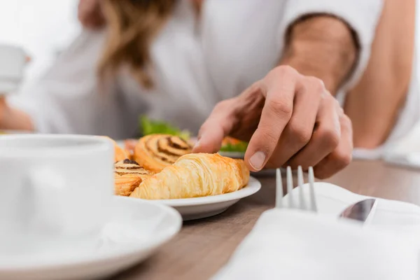 Vista cortada do homem tomando croissant perto da namorada e da xícara em primeiro plano desfocado no hotel — Fotografia de Stock