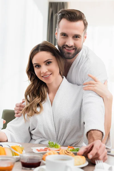 Smiling couple in bathrobes looking at camera near breakfast on blurred foreground on table in hotel — Stock Photo