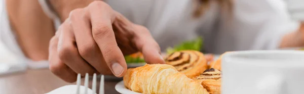 Cropped view of man taking croissant near cup on blurred foreground in hotel, banner — Stock Photo