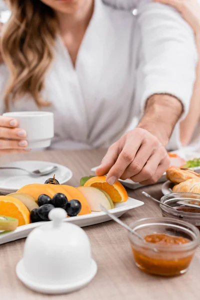 Cropped view of man taking orange slice near food and girlfriend on blurred background in hotel — Stock Photo