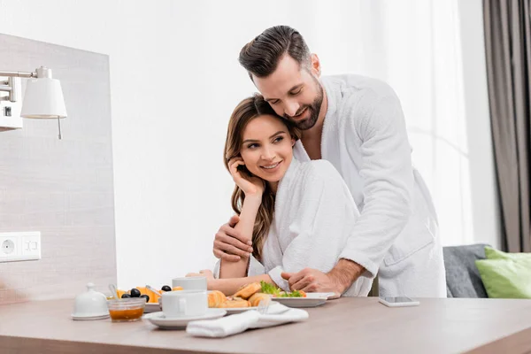 Hombre sonriente en albornoz abrazando a su novia cerca del delicioso desayuno en la habitación del hotel - foto de stock
