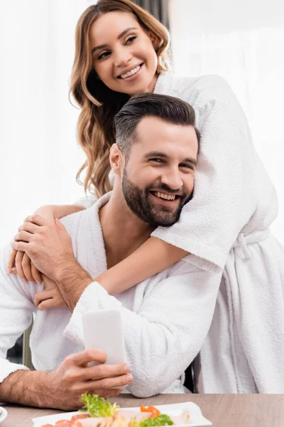 Cheerful woman in bathrobe hugging boyfriend near food on blurred foreground in hotel — Stock Photo