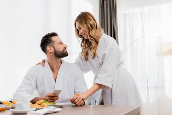 Smiling woman in bathrobe looking at boyfriend with smartphone near breakfast on blurred foreground in hotel — Stock Photo