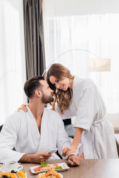 Mujer sonriente abrazando novio en albornoz con teléfono inteligente cerca del desayuno y café en primer plano borroso en la habitación del hotel - foto de stock
