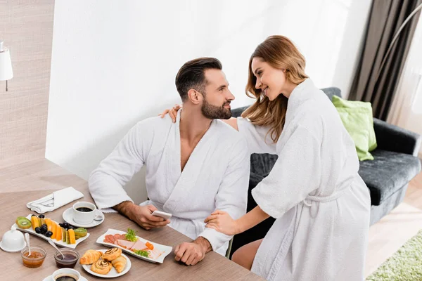 Femme en peignoir embrassant petit ami avec smartphone près de délicieux petit déjeuner et café dans la chambre d'hôtel — Photo de stock